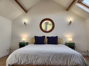 A bedroom with a sloped ceiling, a large bed with white bedding, two blue pillows, two bedside tables with lamps, and a circular window above the bed.