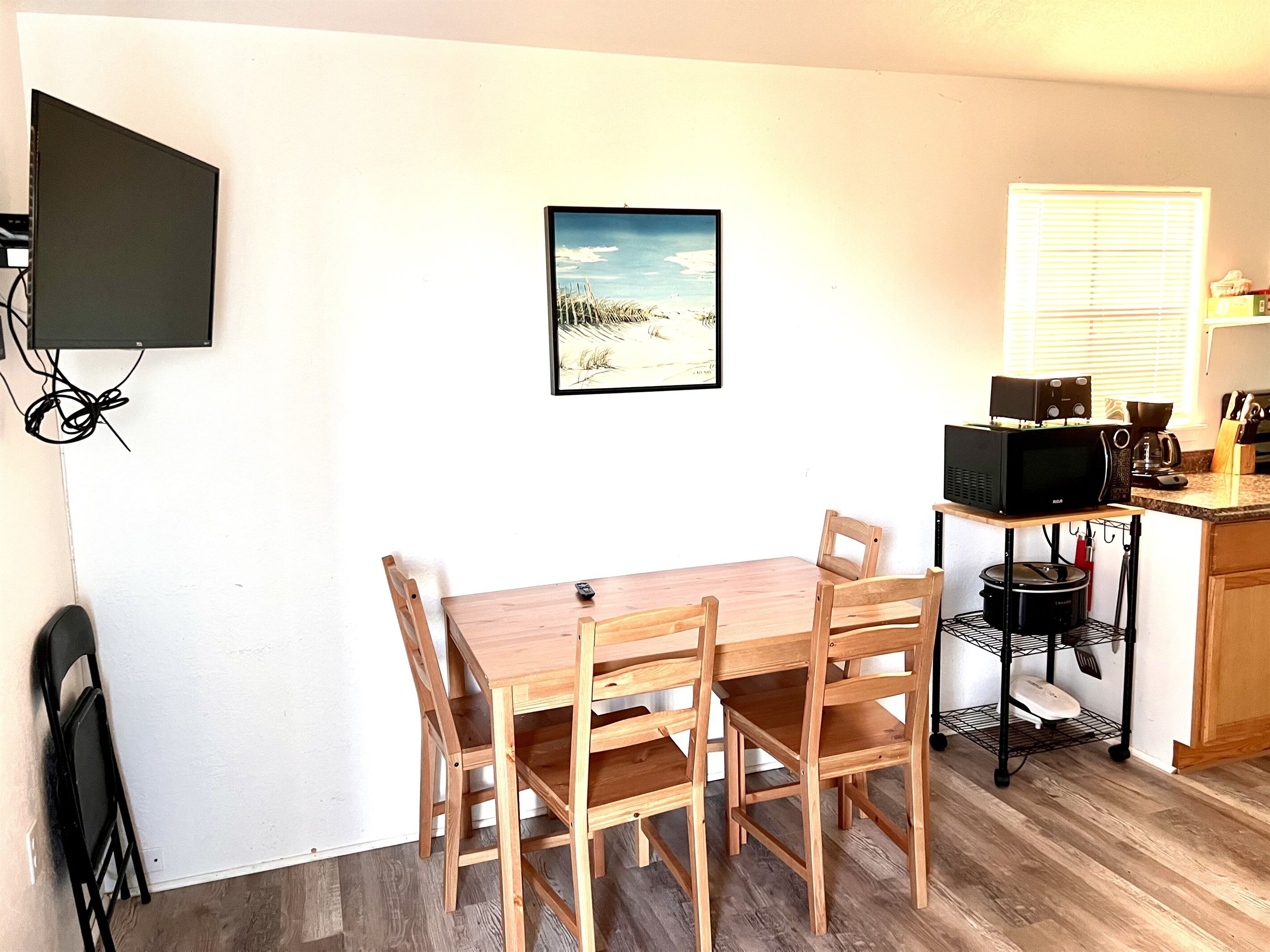 A small kitchen with a wooden table and chairs, a microwave, a coffee maker, and a window with blinds.
