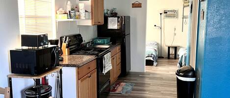 A compact kitchen with wooden cabinets, a black microwave, and a stove. There is a window with blinds, a ceiling fan, and a trash can.