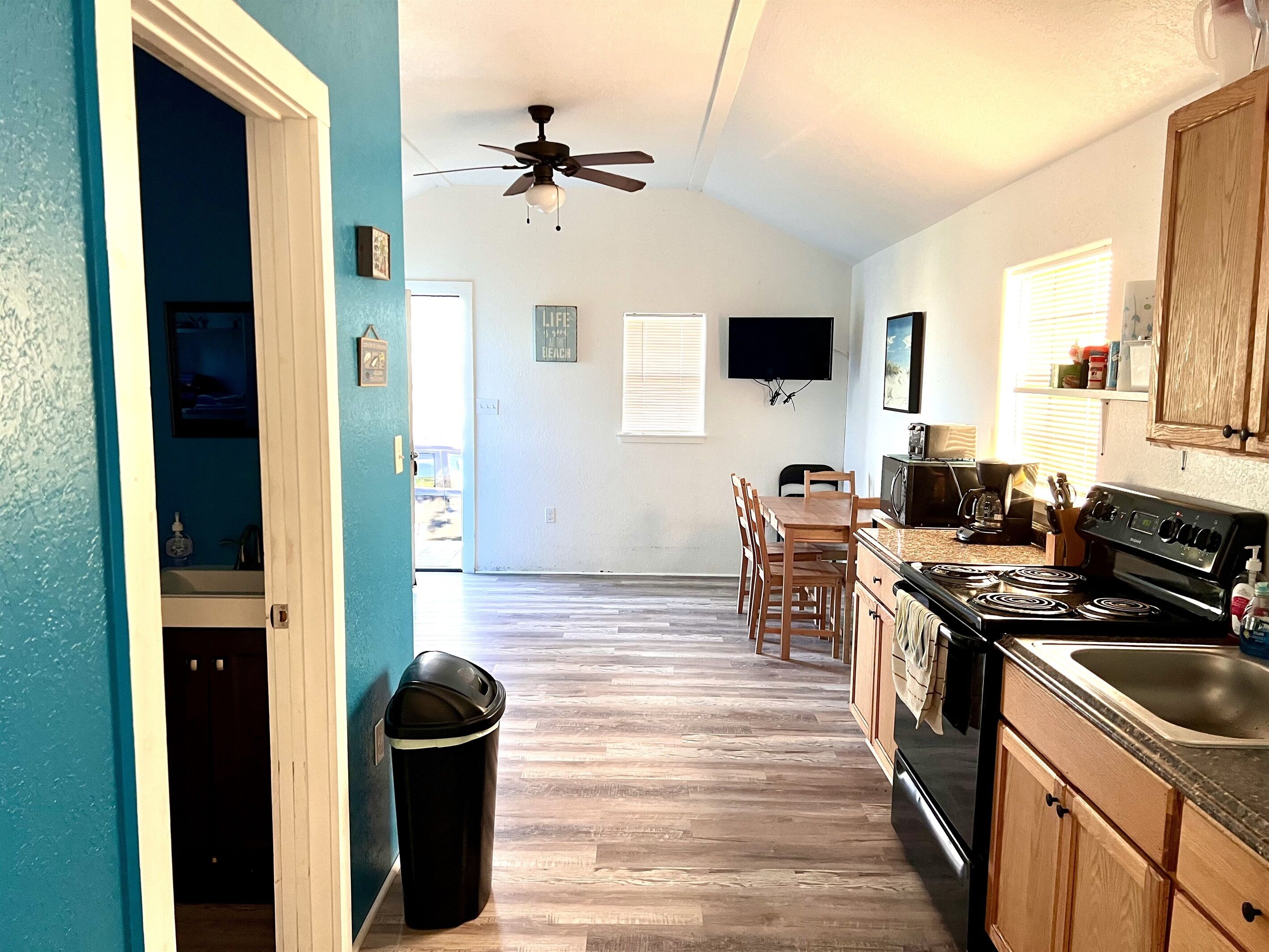 A kitchen with wooden cabinets, a stove, and a microwave. There is a ceiling fan and a trash can.