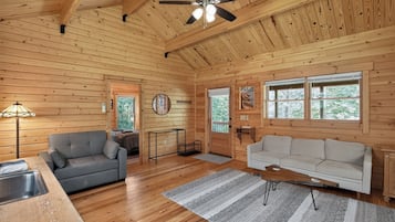A wooden cabin interior with a ceiling fan, two sofas, a dining table, and a window with a view of trees.