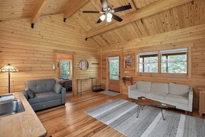 Interior de una cabaña de madera con un ventilador de techo, dos sofás, una mesa de comedor y una ventana con vistas a árboles.