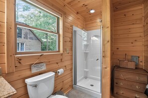 A wooden cabin bathroom with a toilet, a glass shower stall, and a window overlooking a house.