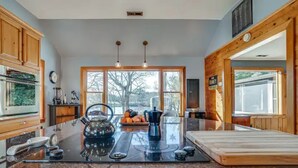 A modern kitchen with wooden cabinets, a large island with a coffee maker, and large windows offering a view of the outdoors.