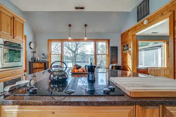 A modern kitchen with wooden cabinets, a large island with a coffee maker, and large windows offering a view of the outdoors. - Pizza Friday (Ridge)