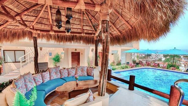 A poolside area with a thatched roof, a curved sofa with patterned cushions, and a view of the pool and surrounding landscape. - San Miguel PV Rentals (Puerto Vallarta)