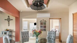 A dining area with a glass table, grey upholstered chairs, and a vase with flowers. There is a cross on the wall and an air conditioning unit. - San Miguel PV Rentals (Puerto Vallarta)