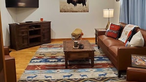 Uma sala de estar com piso de madeira, um sofá marrom, uma mesa de centro, uma televisão, um ventilador de teto e um quadro pendurado na parede.