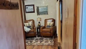 Um corredor com piso de madeira, duas poltronas de couro com almofadas estampadas, uma pequena mesa com abajur e um tapete.