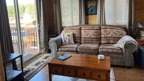 Interior acogedor de una cabaña con un sofá estampado, una mesa de centro y un área de comedor visible a través de ventanas grandes.