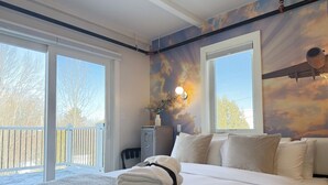 A hotel room with a large bed, a balcony with a view of trees, and a wall with a sunset mural.