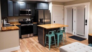 A modern kitchen with dark cabinets, stainless steel appliances, a wooden countertop, and a bar area with two chairs. - Charming Downtown Cody Cottage (#1) (Cody)