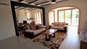 Una sala de estar con sofá, sillones, una mesa de centro y televisor. Hay ventanas y un arco de entrada.