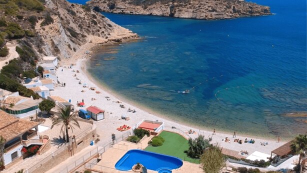 Una playa costera con aguas azul cristalina, una orilla arenosa y un pequeño bote. Se observan edificios con balcones y una piscina.