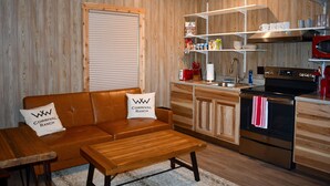 A cozy living room with a brown leather sofa, a wooden coffee table, and a kitchen area with stainless steel appliances and wooden cabinetry. - Charming Cabin Retreat in Texas Hill Country (Boerne)