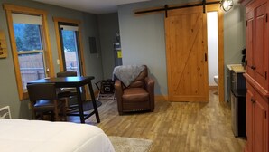 A cozy room with a bed, a dining table with chairs, a leather chair, a small table with a blanket, a wooden door, and a kitchen area with cabinets and a sink.
