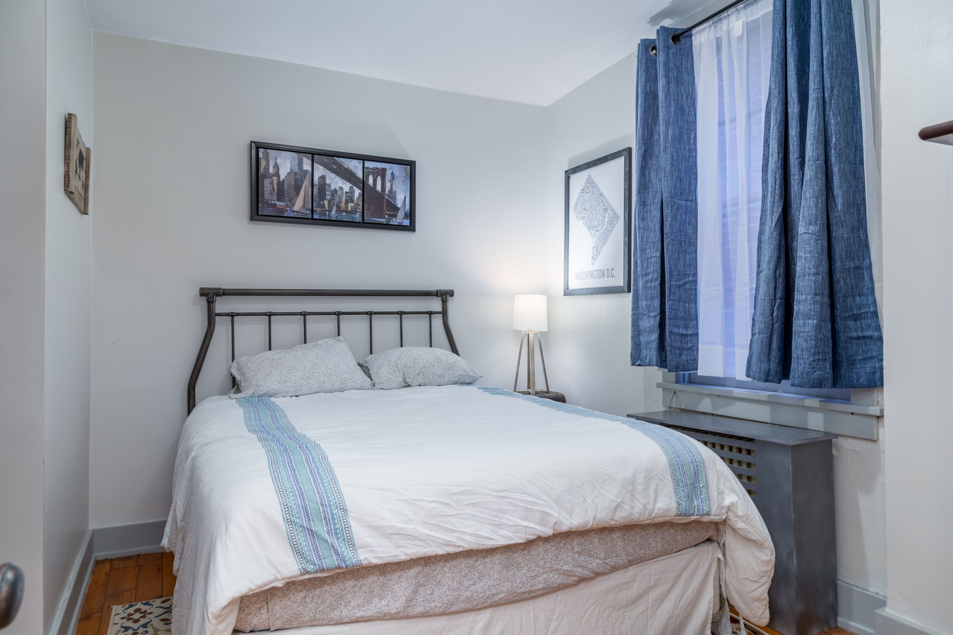 A neatly made bed with a white and blue striped blanket, a framed cityscape picture on the wall, and a window with blue curtains.