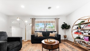 A living room with a black sofa, a round coffee table, a patterned rug, a bookshelf, and a window with curtains. - The Gathering Place - Boho Getaway Perfect For Family Stays! (Lubbock)