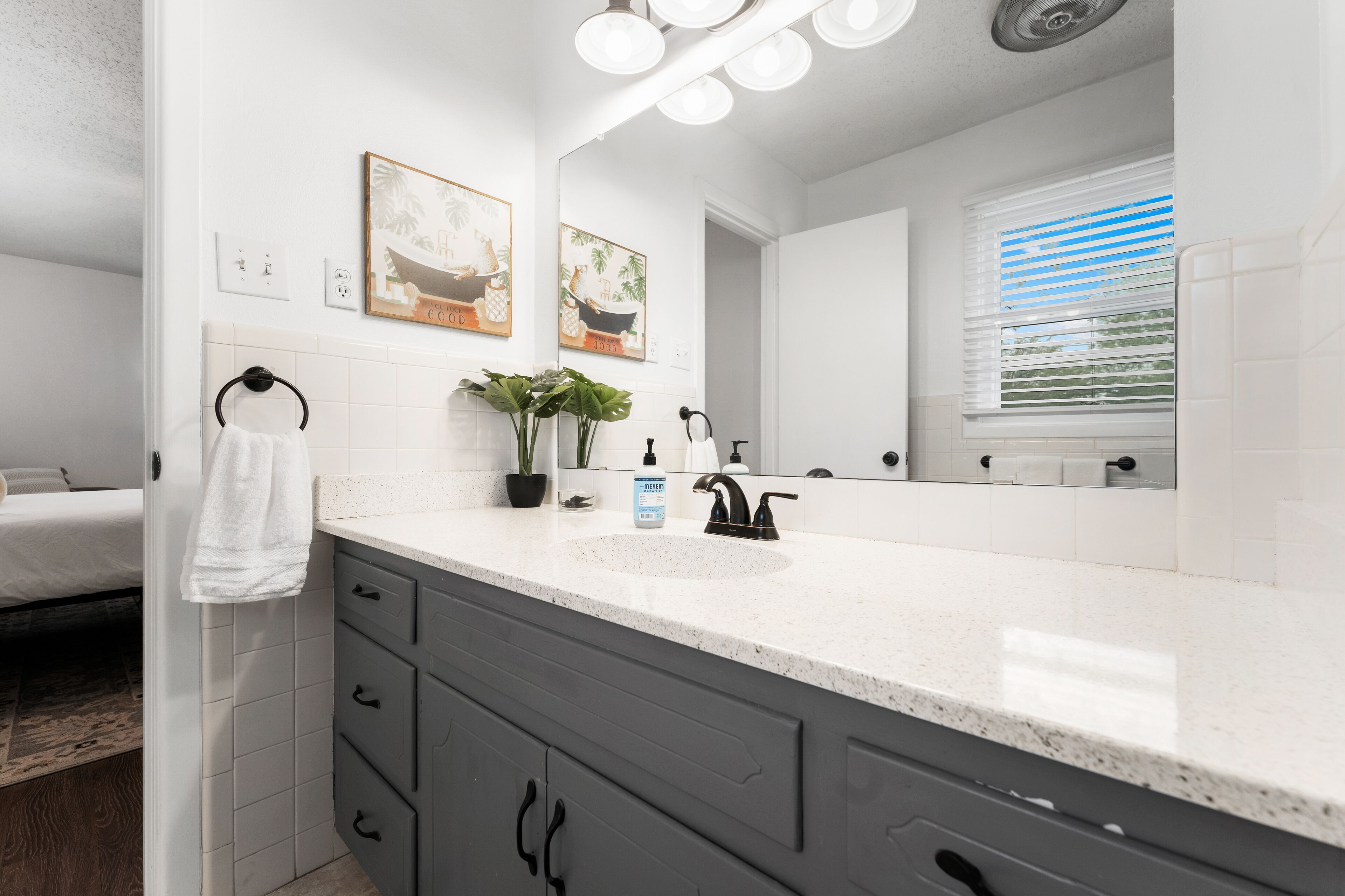 A modern bathroom with a white sink, dark grey cabinets, and a large mirror. There are two framed pictures on the wall, a towel rack, and a window with blinds.