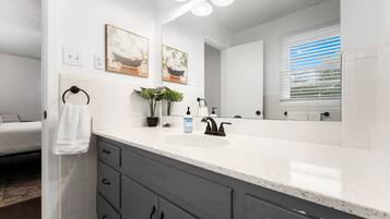 A modern bathroom with a white sink, dark grey cabinets, and a large mirror. There are two framed pictures on the wall, a towel rack, and a window with blinds.
