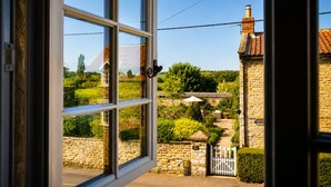 Blick durch ein Fenster auf ein Steinhaus mit Garten, weißem Zaun und einem Ziegelkamin.