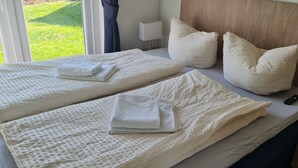 Habitación de hotel con dos camas, cada una con colcha blanca texturizada y almohadas, una mesita de noche con lámpara, y vistas al exterior a través de una ventana.
