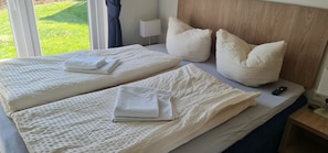 Two beds with white mattresses and a nightstand with a lamp and remote control.