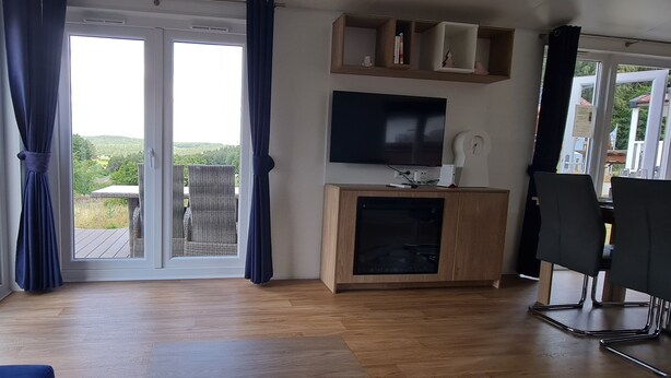 Una habitación con un gran ventanal corredero que da a un balcón, un televisor de pantalla plana montado en la pared y un mueble de madera con un televisor encima.