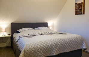 Ein ordentlich bezogenes Bett mit gesteppter Decke und Kissen, eine Nachttischlampe und ein an der Wand befestigter Bilderrahmen.