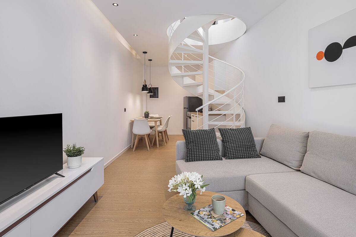 Una sala moderna con un televisor de pantalla plana, un sofá, una mesa de centro con una revista y flores, y una escalera de caracol al fondo.