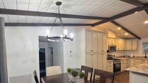 Un comedor amplio con mesa y sillas de madera, una lámpara de araña y vistas al área de la cocina con armarios blancos y un microondas.