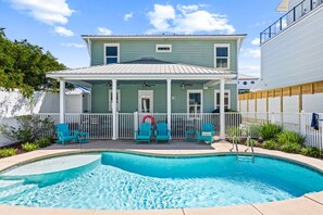 A pool area with a house in the background, a covered patio with chairs, and a fence.