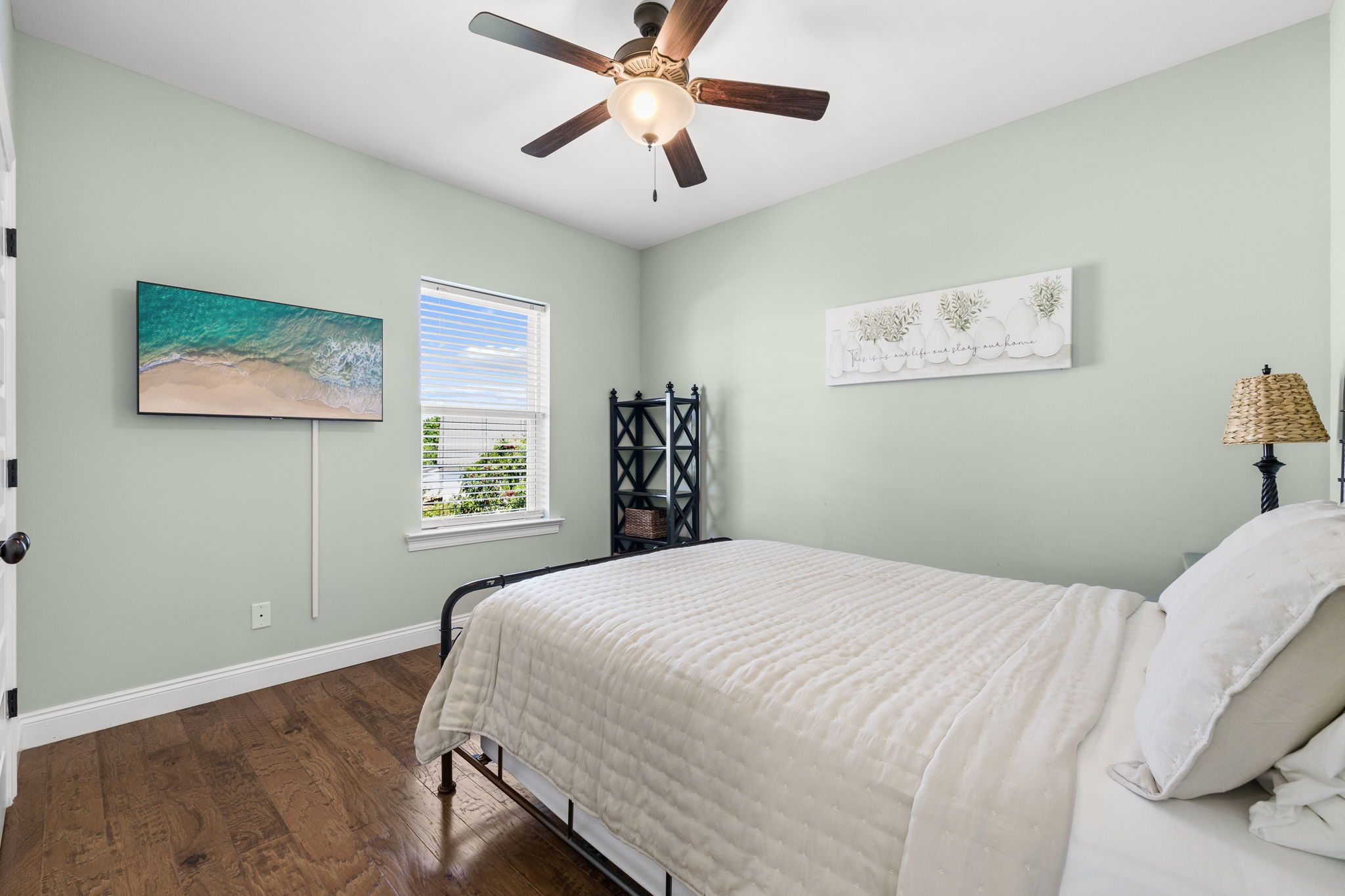 A bedroom with a bed, a ceiling fan, a window with blinds, a wall-mounted shelf, and a painting.