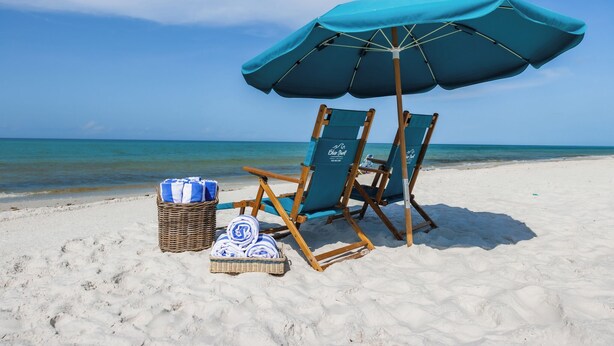Eine Strandansicht mit zwei Holzliegen, einem blauen Sonnenschirm und einem Korbwäschekorb mit Handtüchern.