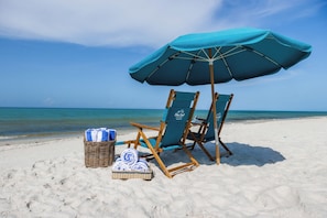 A beach scene with two wooden beach chairs, a blue umbrella, and a wicker basket with towels. - 5 Bed, 5 Bath Beach Home/only a 3 Minute Walk To The Beach! Sleeps 12 (Panama City Beach)