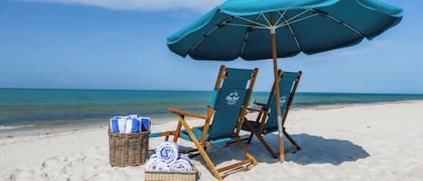 A beach scene with two wooden beach chairs, a blue umbrella, and a wicker basket with towels.