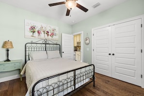 Une chambre à coucher comprenant un lit, un ventilateur de plafond, une table de chevet, une lampe, un tableau accroché au mur et une porte.