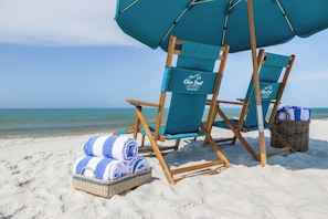 A beach setting with two blue lounge chairs, a wicker basket with towels, and a large umbrella. - 5 Bed, 5 Bath Beach Home/only a 3 Minute Walk To The Beach! Sleeps 12 (Panama City Beach)