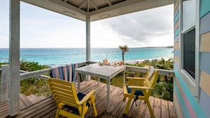 A wooden deck with a table and chairs, overlooking a beach with the ocean and a cloudy sky. - Cayo Loco 1 Bedroom Home (Governor's Harbour)
