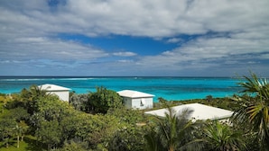 A coastal view with two white buildings surrounded by lush greenery and a turquoise sea under a partly cloudy sky. - Cayo Loco 1 Bedroom Home (Governor's Harbour)