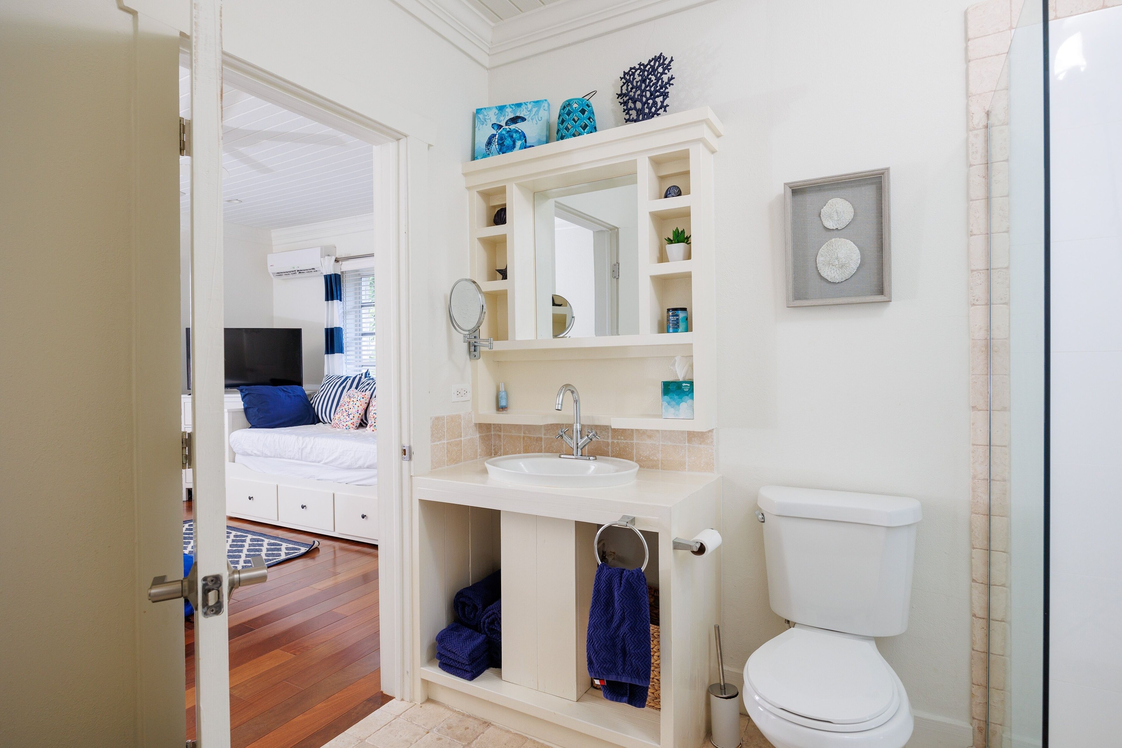 A bathroom with a white sink, mirror, and toilet. There is a blue towel hanging on the door, and a shelf with decorative items above the sink.