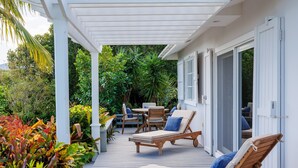 A covered outdoor area with wooden lounge chairs and a table, surrounded by lush greenery. - One Palm Hill 3 Bedroom Villa (Governor's Harbour)