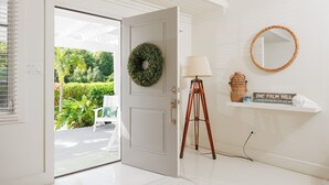 A white door leading to a patio with a white chair, a lamp, a mirror, and a shelf with a book and a basket. - One Palm Hill 3 Bedroom Villa (Governor's Harbour)