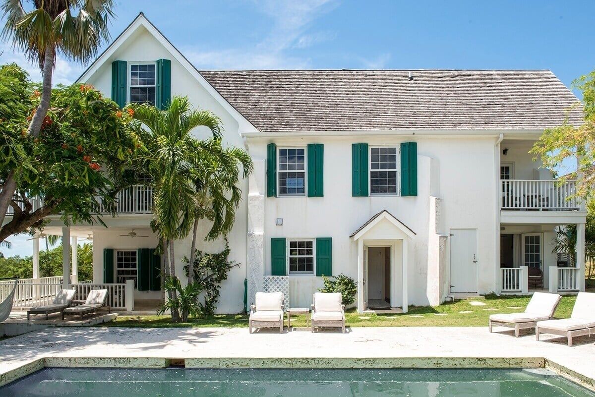 A white building with green shutters, a pool, and outdoor seating.
