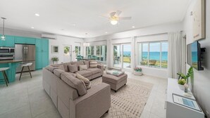 Una sala de estar moderna con un sofá seccional color beige, una cocina con gabinetes verde mar y un gran ventanal corredizo que da a un balcón con vistas al océano.