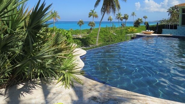 A swimming pool with clear blue water, surrounded by lush greenery and palm trees, with a view of the ocean and a building in the background.