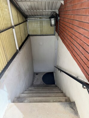 A concrete stairway with a metal handrail, leading to a door with a metal frame and a white wall on one side and a red brick wall on the other. - Cozy Basement Retreat • King Bed • Near TRAX (South Salt Lake)