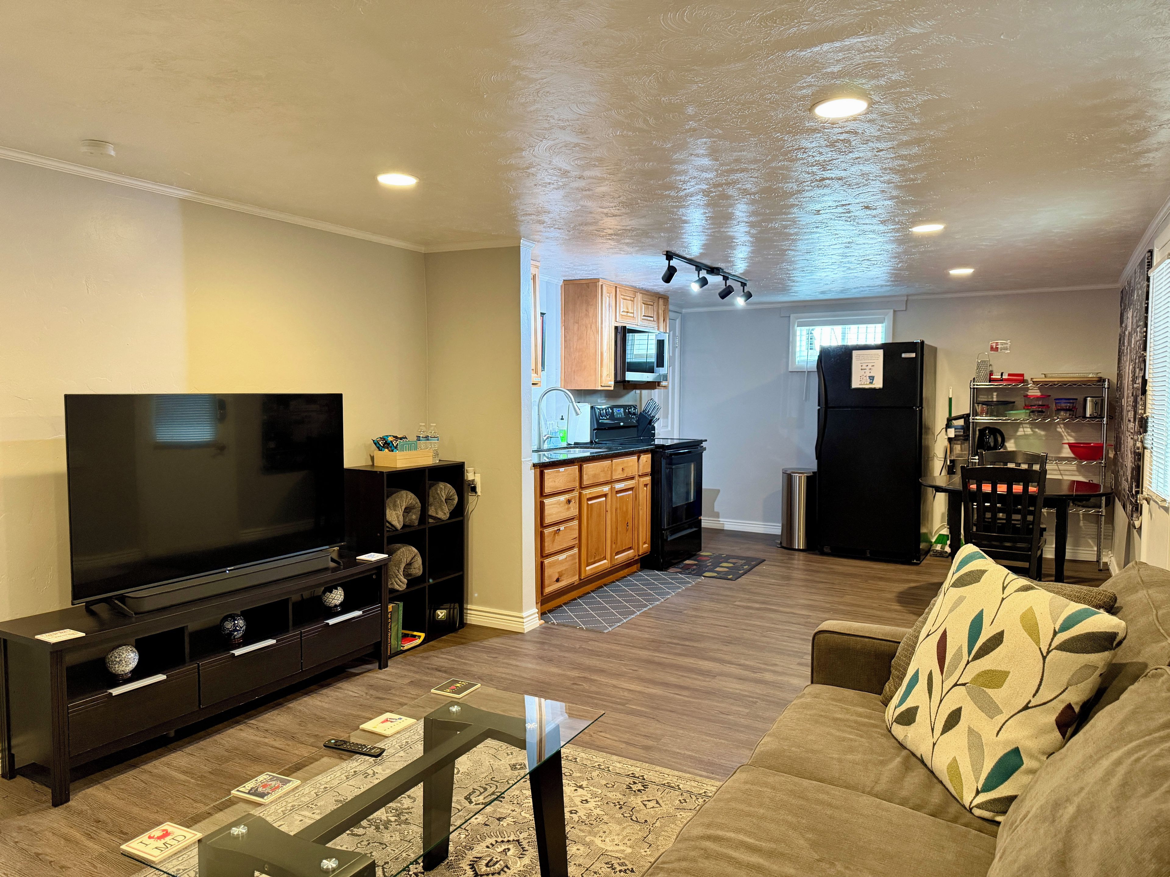 A living room with a flat-screen TV, wooden cabinets, a kitchen area with a microwave, stove, and refrigerator, and a sofa with patterned cushions.