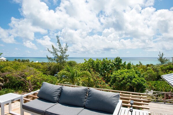 Une terrasse en bois avec un canapé gris et des coussins, donnant sur un paysage tropical luxuriant et offrant une vue sur l’océan.