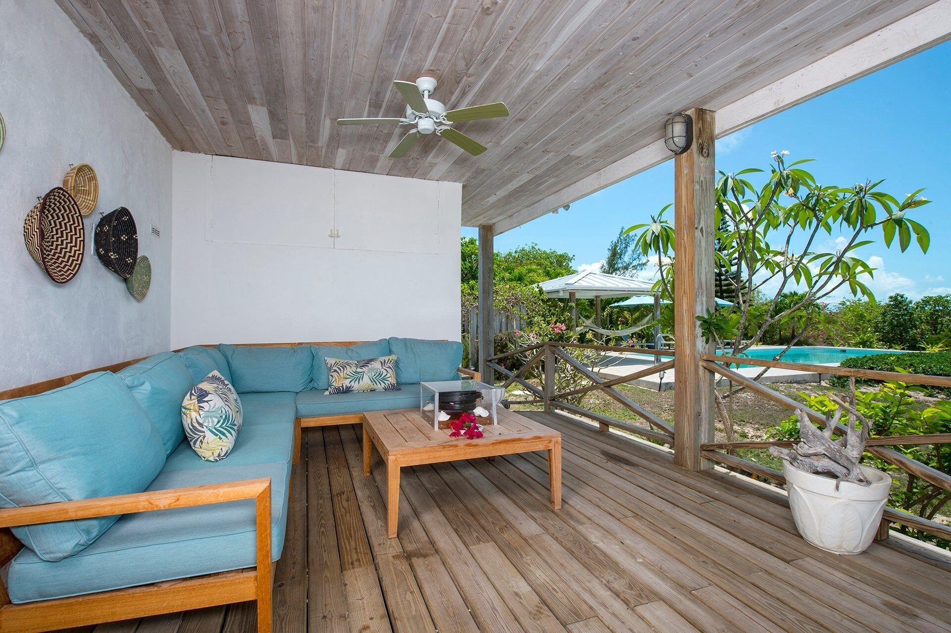 Ein überdachter Außenbereich mit einer hölzernen Bank und einem blauen Sofa, einem Deckenventilator und Blick auf Pool und Grünflächen.